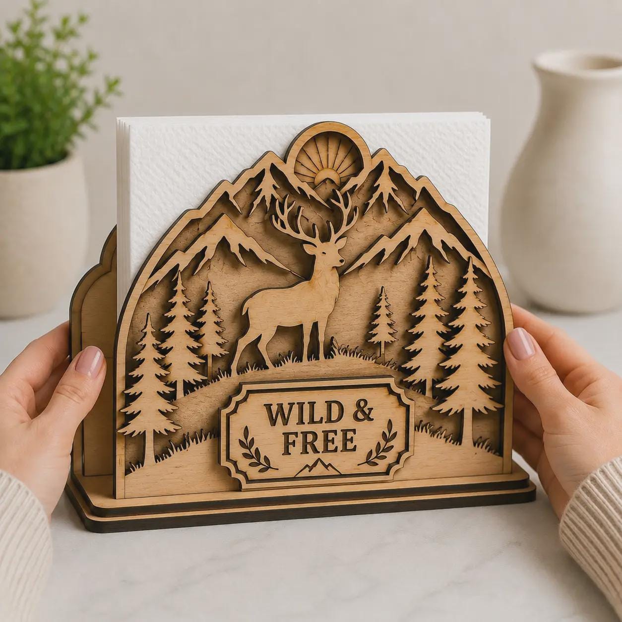 Rustic deer napkin holder displayed on a wooden table, featuring layered mountain scenery, woodland elements, pine trees, and decorative laser cut craftsmanship.